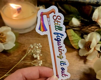 Può includere: Un adesivo raffigurante un bastone da passeggio a fumetti con un nappa arcobaleno e la scritta "Still figuring it out". Il bastone ha un'impugnatura marrone, un fusto argentato e una punta viola. Il testo è in gradiente arcobaleno.