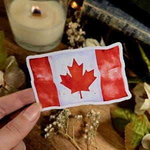 Puede incluir: Una pegatina de acuarela de la bandera canadiense. La bandera es roja y blanca con una hoja de arce roja en el centro. La pegatina est&aacute; sobre una superficie de madera.