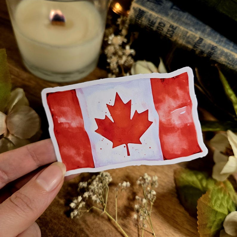 Puede incluir: Una pegatina de acuarela de la bandera canadiense. La bandera es roja y blanca con una hoja de arce roja en el centro. La pegatina est&aacute; sobre una superficie de madera.