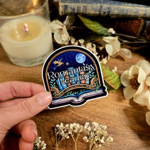 May include: A sticker with the text "Romantasy Reader" in a decorative font, featuring a night sky with a moon, stars, and a dragon. The sticker has a black border and is set against a background of books and flowers.