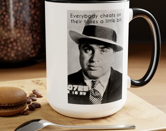 May include: White ceramic mug with a black interior and handle. The mug features a black and white image of a man in a hat with the text "Everybody cheats on their taxes a little bit."