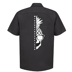 Può includere: Una camicia da lavoro nera a maniche corte con una grafica di teschio di ananas bianco e il testo "WIRED PINEAPPLE CO" sul retro.