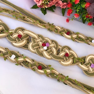May include: Decorative rope trim in shades of green, beige, and brown, adorned with small red, white, and purple fabric roses and green leaves. The trim is intricately woven and appears to be handmade, suitable for various craft projects.