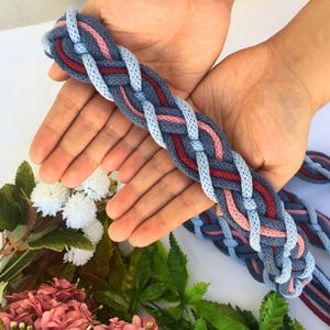 May include: Braided textile belt in shades of blue, red, and pink. The belt is held in open hands against a white background, with floral accents in the lower left corner. The belt's woven design is visible.