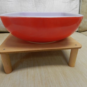 Puede incluir: Un tazón de cerámica rojo y blanco sobre un soporte de madera. El tazón es redondo y tiene un acabado liso y brillante. El soporte está hecho de madera natural y tiene tres patas.