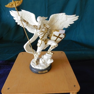 Puede incluir: Una figura blanca y dorada de un ángel con grandes alas, sosteniendo un báculo con un pájaro dorado y un escudo. El ángel está de pie sobre una base en forma de nube. La figura está sobre una mesa de madera.