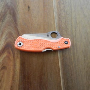 Puede incluir: Una navaja plegable naranja con un mango texturizado y una hoja plateada. La navaja está cerrada y descansa sobre una superficie de madera. La hoja tiene un agujero circular cerca del mango.