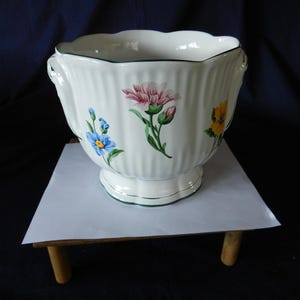 Puede incluir: Una maceta de cerámica blanca con borde festoneado y textura acanalada vertical. El exterior presenta motivos florales pintados a mano, incluyendo flores azules, rosas y amarillas con tallos verdes. La base es de pedestal con un borde verde oscuro.