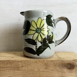 Pode incluir: Um jarro de cerâmica cinza com um design floral. O jarro tem uma flor amarela com um centro azul e folhas verdes. O jarro tem uma alça marrom.