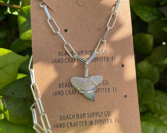 May include: Silver chain necklace with a shark tooth pendant. The pendant is wrapped in silver wire and has a blue-grey color. The tag below the pendant reads "Beach Bum Supply Co. Hand Crafted in Jupiter FL."