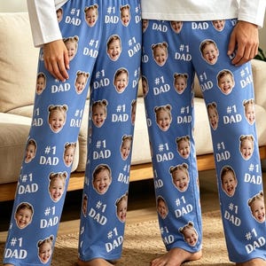 May include: Two pairs of blue pajama pants featuring a repeating pattern of a child's face and the text #1 DAD in white. The pants are worn with plain white long-sleeved shirts. The fabric is light blue and the print is evenly distributed throughout.