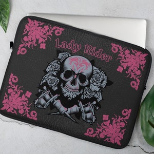 May include: Black laptop sleeve featuring a skull design with roses and tattoo machine illustrations. The text "Lady Rider" is displayed in pink. The sleeve has a textured appearance and a zipper closure.