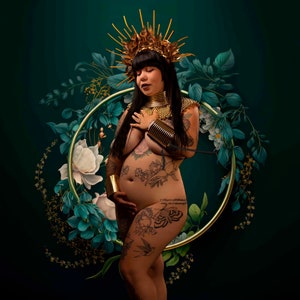 May include: A pregnant person with tattoos and a gold crown stands in front of a green background with a gold circle and floral designs. The person is wearing a gold necklace and bracelets.