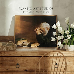 Op de afbeelding: Een ingelijst schilderij van een man die bidt met zijn hoofd gebogen. Het schilderij staat op een houten tafel met een vaas met witte tulpen en een rieten mand.