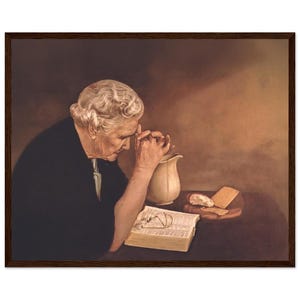 Op de afbeelding: Een vrouw met grijs haar bidt met haar handen gevouwen. Ze draagt een zwarte jurk en zit aan een tafel met een boek, een kan en een stuk brood.