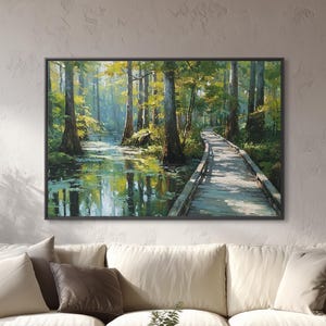 May include: A framed landscape painting depicts a serene forest scene with a wooden boardwalk winding through a swamp. The artwork features lush green trees, a reflective body of water, and a bright, sunny sky. The painting is displayed above a cream-colored sofa.