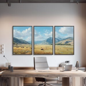 May include: Three framed landscape paintings depicting a sunny meadow with mountains and a blue sky. The paintings hang above a wooden desk in a modern office setting. A wooden bookshelf and a gray office chair are also visible.