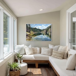 May include: A framed landscape painting of a lake surrounded by trees and rocks hangs on a wall. The room features a built-in seating area with white cushions and decorative pillows. Potted plants and a small stool are in the foreground.
