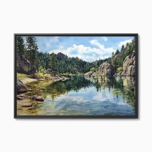 May include: A framed landscape painting depicts a serene lake scene. The artwork features a calm, reflective lake surrounded by lush green trees, large rock formations, and a bright blue sky with fluffy white clouds. The painting is encased in a black frame.