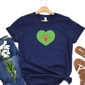 May include: A navy blue t-shirt with a green heart featuring a red frog print. The shirt is displayed on a white background with a pair of brown sandals and blue jeans.