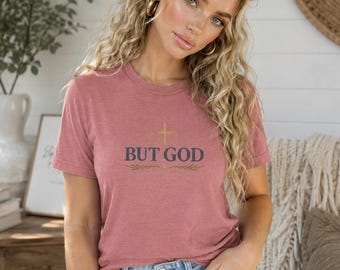 May include: A dusty rose short-sleeved t-shirt featuring the text BUT GOD in dark blue lettering, centered above a small gold cross graphic and decorative leaf accents. The shirt is paired with light blue denim jeans with a visible metal button.