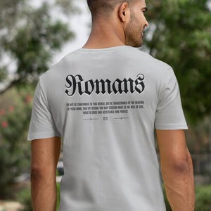 May include: A pale grey t-shirt featuring the word "Romans" in a gothic font and a Bible verse on the back. The short-sleeved shirt is worn by a person outdoors.