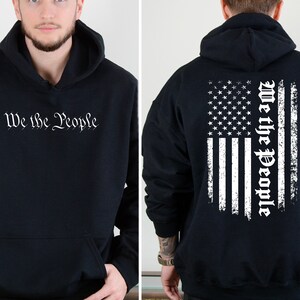 K&ouml;nnte beinhalten: Schwarzer Kapuzenpullover mit einem wei&szlig;en, abgenutzten amerikanischen Flaggenmuster auf der R&uuml;ckseite und dem Text "We the People" in einer Schriftart auf der Vorderseite.