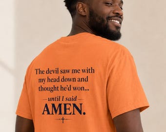 May include: An orange short-sleeved t-shirt featuring black text on the back that reads: The devil saw me with my head down and thought he'd won... until I said AMEN. A small cross symbol is printed below the word AMEN on the back of the shirt.