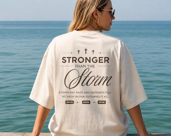 May include: An off-white short-sleeve t-shirt featuring dark brown text on the back that reads STRONGER THAN THE STORM, followed by the phrase STORMS MAY RAGE AND DARKNESS FALL YET FAITH WITHIN OUTSHINES IT ALL, and the words FAITH OVER FEAR.