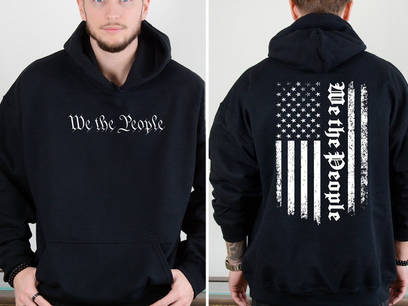 K&ouml;nnte beinhalten: Schwarzer Kapuzenpullover mit einem wei&szlig;en, abgenutzten amerikanischen Flaggenmuster auf der R&uuml;ckseite und dem Text "We the People" in einer Schriftart auf der Vorderseite.