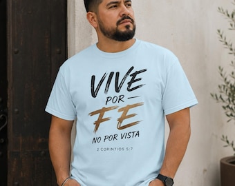 May include: A light blue short-sleeve t-shirt featuring the black and gold brushstroke text "VIVE POR FE NO POR VISTA" and "2 CORINTIOS 5:7" printed on the front. The shirt has a crew neckline and a standard fit, showcasing a clean design.
