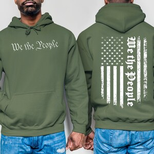 K&ouml;nnte beinhalten: Ein gr&uuml;ner Kapuzenpullover mit dem Aufdruck "We the People" auf der Vorderseite und einem abgenutzten amerikanischen Flaggenmuster auf der R&uuml;ckseite.