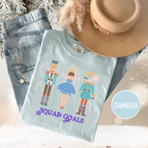 May include: A light blue t-shirt with a graphic of a nutcracker, a ballerina, and a mouse in a nutcracker costume. The text "Squad Goals" is printed below the graphic. The shirt is made of Chambray fabric.