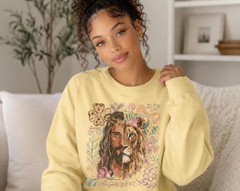 May include: A pale yellow crewneck sweatshirt featuring a central graphic print that depicts a split face, with one side showing a human figure with long brown hair and a crown of thorns, and the other side showing a lion, surrounded by floral motifs.