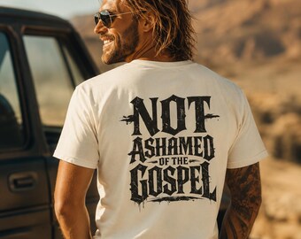 May include: A cream-colored short-sleeve t-shirt featuring black distressed-style lettering on the back that reads "NOT ASHAMED OF THE GOSPEL" in a bold, stylized font. The shirt has a crew neckline and a standard fit, showcasing a clean design.