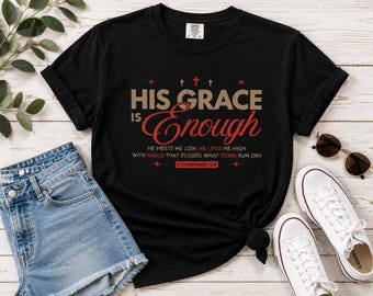 May include: A black short-sleeve t-shirt featuring the text 'HIS GRACE IS ENOUGH' in tan and red, with additional smaller text reading 'HE MEETS ME LOW, HE LIFTS ME HIGH WITH GRACE THAT FLOODS WHAT TEARS RUN DRY' and '2 CORINTHIANS 12:9'.