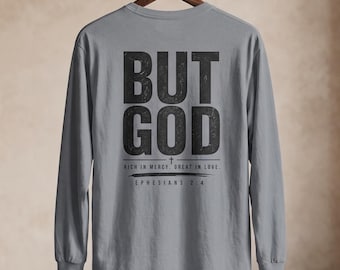 May include: A grey long-sleeve t-shirt featuring black text on the back that reads BUT GOD, RICH IN MERCY, GREAT IN LOVE, and EPHESIANS 2:4. The shirt is displayed on a wooden hanger, showcasing a crew neckline and long sleeves with ribbed cuffs.