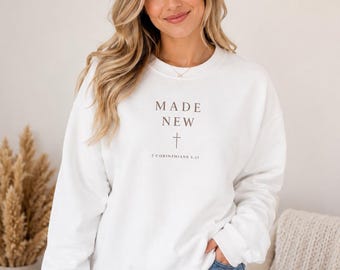 May include: A white crewneck sweatshirt featuring the text MADE NEW in dark brown lettering, centered above a small cross symbol and the scripture reference 2 CORINTHIANS 5:17. The garment has long sleeves with ribbed cuffs and a ribbed hemline.