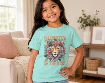 May include: A light turquoise short-sleeve t-shirt featuring a graphic print of a lion's head surrounded by colorful flowers. The text "DAUGHTER OF THE KING" is printed in a bold, orange-toned font above and below the lion illustration.
