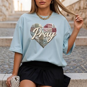May include: Light blue t-shirt featuring a heart-shaped design with a patchwork pattern and the word "Pray" in a script font. The shirt has short sleeves and a relaxed fit, suitable for casual wear.