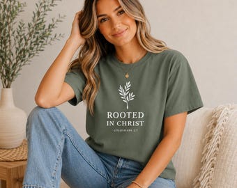 May include: An olive green short-sleeve t-shirt featuring a white graphic of a leafy sprig above the text "ROOTED IN CHRIST" and "COLOSSIANS 2:7" in a clean, sans-serif font. The shirt is paired with light blue denim jeans.