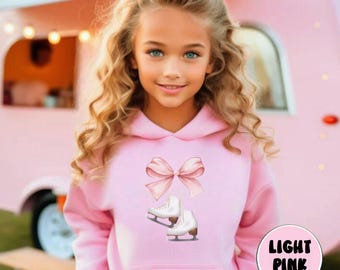 May include: A young girl wearing a light pink hoodie with a pink bow and white ice skates printed on the front. The hoodie has a hood and a kangaroo pocket. The girl has long blonde hair.