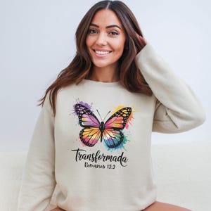 May include: Beige sweatshirt with a vibrant butterfly design. The butterfly is depicted with a watercolor effect, showcasing a spectrum of colours. The word "Transformada" and the text "Romanos 12:2" are printed below the butterfly.