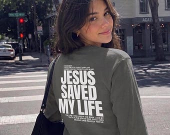 May include: An olive green long-sleeve shirt featuring white text on the back that reads JESUS SAVED MY LIFE, along with smaller printed verses above and below the main phrase. The shirt is paired with a black shoulder bag and dark denim jeans.