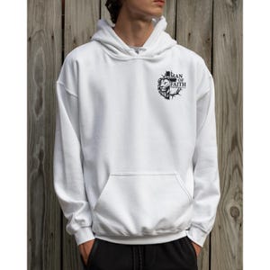 May include: White hoodie with a front pocket and a hood. The hoodie features a black graphic on the upper chest that includes a lion's head and the words "MAN OF FAITH". The background is a wooden wall.