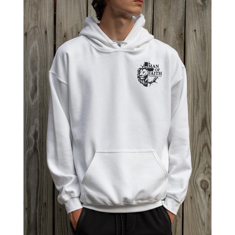 May include: White hoodie with a front pocket and a hood. The hoodie features a black graphic on the upper chest that includes a lion's head and the words "MAN OF FAITH". The background is a wooden wall.