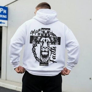 May include: White hoodie with a black graphic on the back. The graphic features a lion's face within a cross, with the words "Man of Faith" and "1 Corinthians 16:13". The hoodie has a hood and long sleeves.