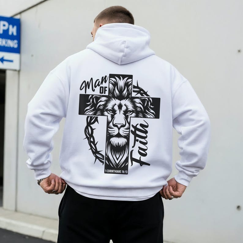 May include: White hoodie with a black graphic on the back. The graphic features a lion's face within a cross, with the words "Man of Faith" and "1 Corinthians 16:13". The hoodie has a hood and long sleeves.