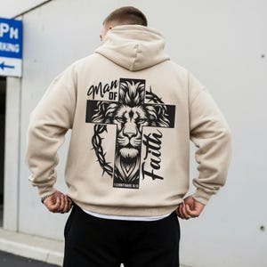 May include: Beige hoodie with a large graphic on the back. The graphic features a lion's face within a cross, with the words "Man of Faith" and a Bible verse. The hoodie has a hood and long sleeves.
