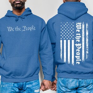 K&ouml;nnte beinhalten: Zwei blaue Kapuzenpullover mit dem Aufdruck "We the People". Ein Pullover hat den Aufdruck auf der Vorderseite, der andere eine abgenutzte amerikanische Flagge mit dem Aufdruck auf der R&uuml;ckseite.